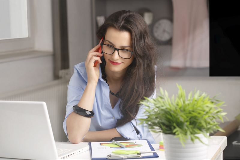 A woman with glasses is talking on the phone while working on a laptop at a desk with papers and a small plant.