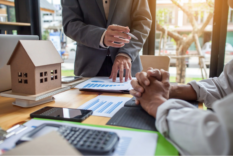 Two professionals having a discussion in an office with charts, documents, a calculator, and a small house model on the table.