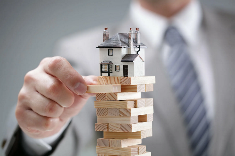 A person in a suit carefully placing a small house model on a precarious stack of wooden blocks.