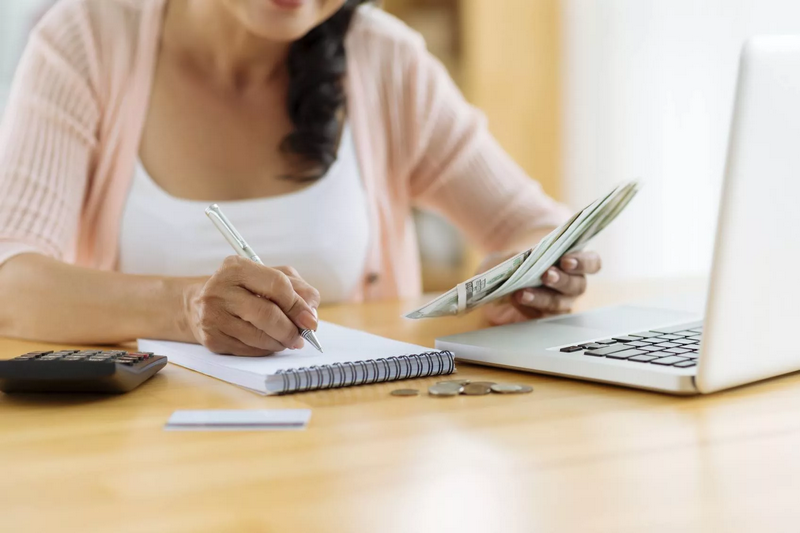 A person calculates finances at a desk using a notebook, calculator, and laptop.