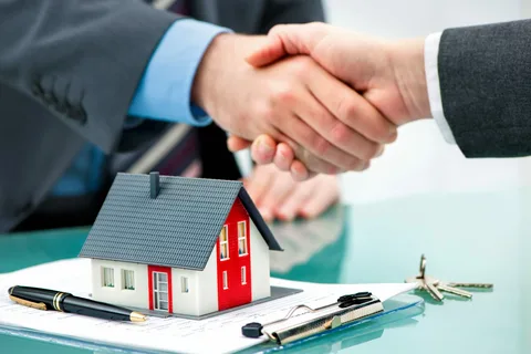 Two individuals shaking hands over a small model house, documents, and keys on a table.