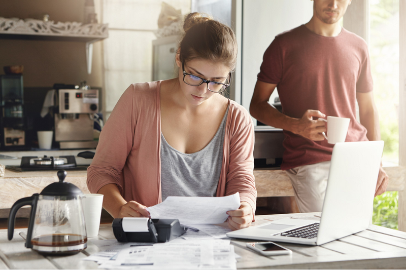 A woman is working on finances at a table with papers and a laptop, while a man stands nearby drinking coffee.