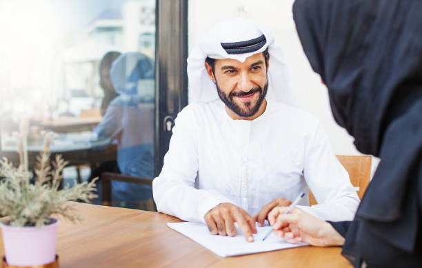 Two people in traditional attire discuss real estate documents in a Dubai office setting.