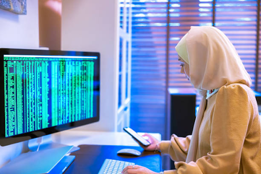 A woman wearing a hijab smiles while holding a credit card and using a smartphone next to a laptop.